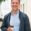 Cheerful guy enjoying outdoor coffee break. Young Caucasian man standing in city street, holding paper cup and smiling at camera. Takeaway coffee concept